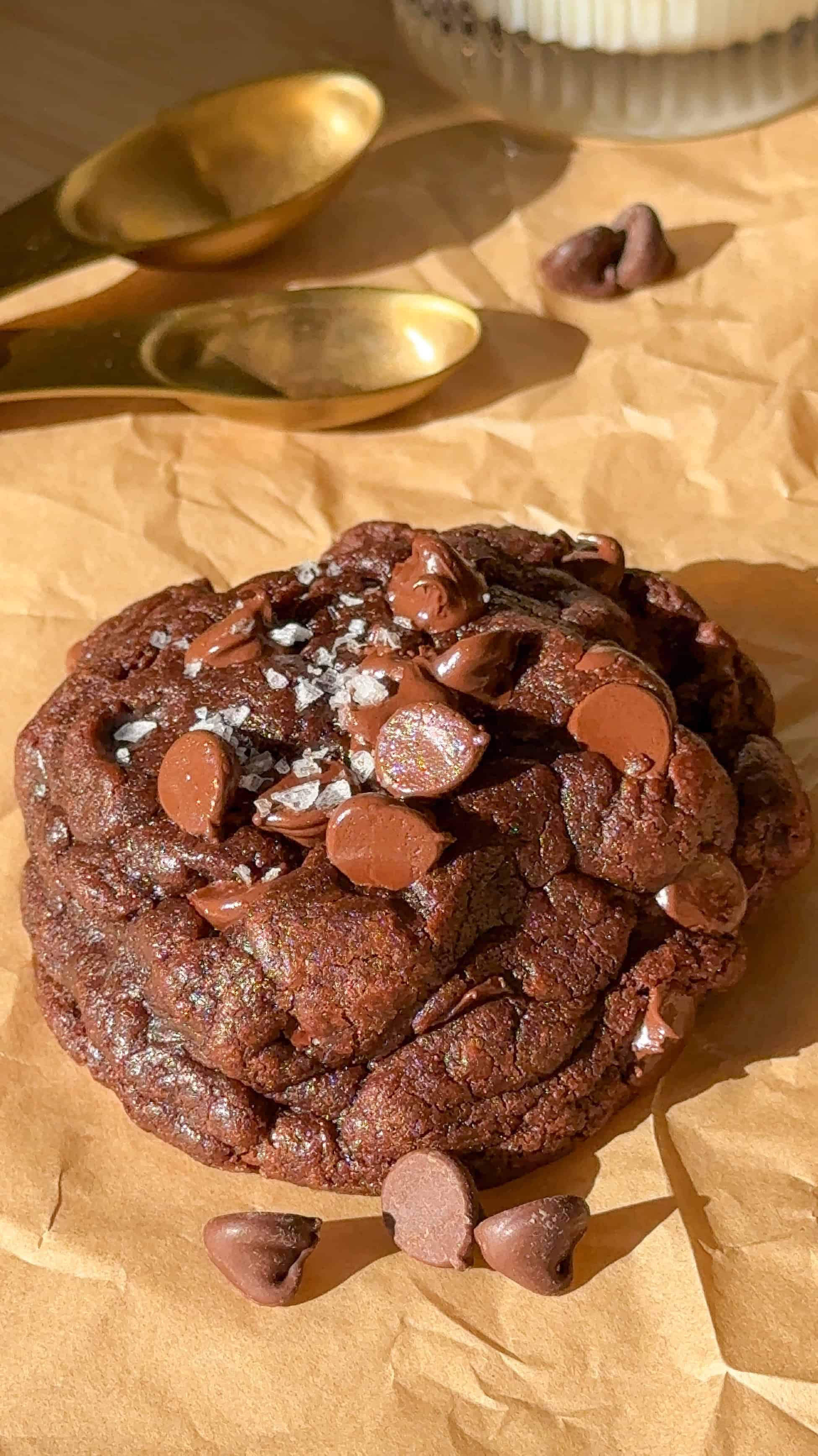 two of my single serve double chocolate chip cookies on a table with a glass of milk, chocolate chips, sea salt, and measuring cups.