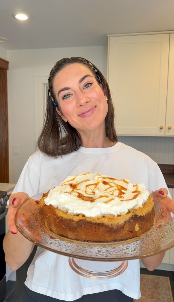 Mallory holding a pumpkin cheesecake with a Biscoff crust, topped with whipped cream and a salted caramel drizzle
